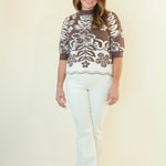 A woman modeling a brown and white patterned mock-neck top with elbow-length sleeves, paired with white pants, standing against a plain cream background.