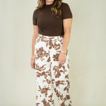 A woman modeling a brown short-sleeve mock neck top and high-waisted, wide-leg pants with a brown and white cow print, standing against a solid light beige studio background.