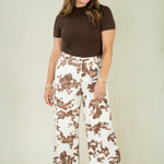 A woman modeling a brown short-sleeve mock neck top and high-waisted white wide-leg pants with a brown abstract print, paired with brown leather cowboy boots, against a plain light beige studio background.