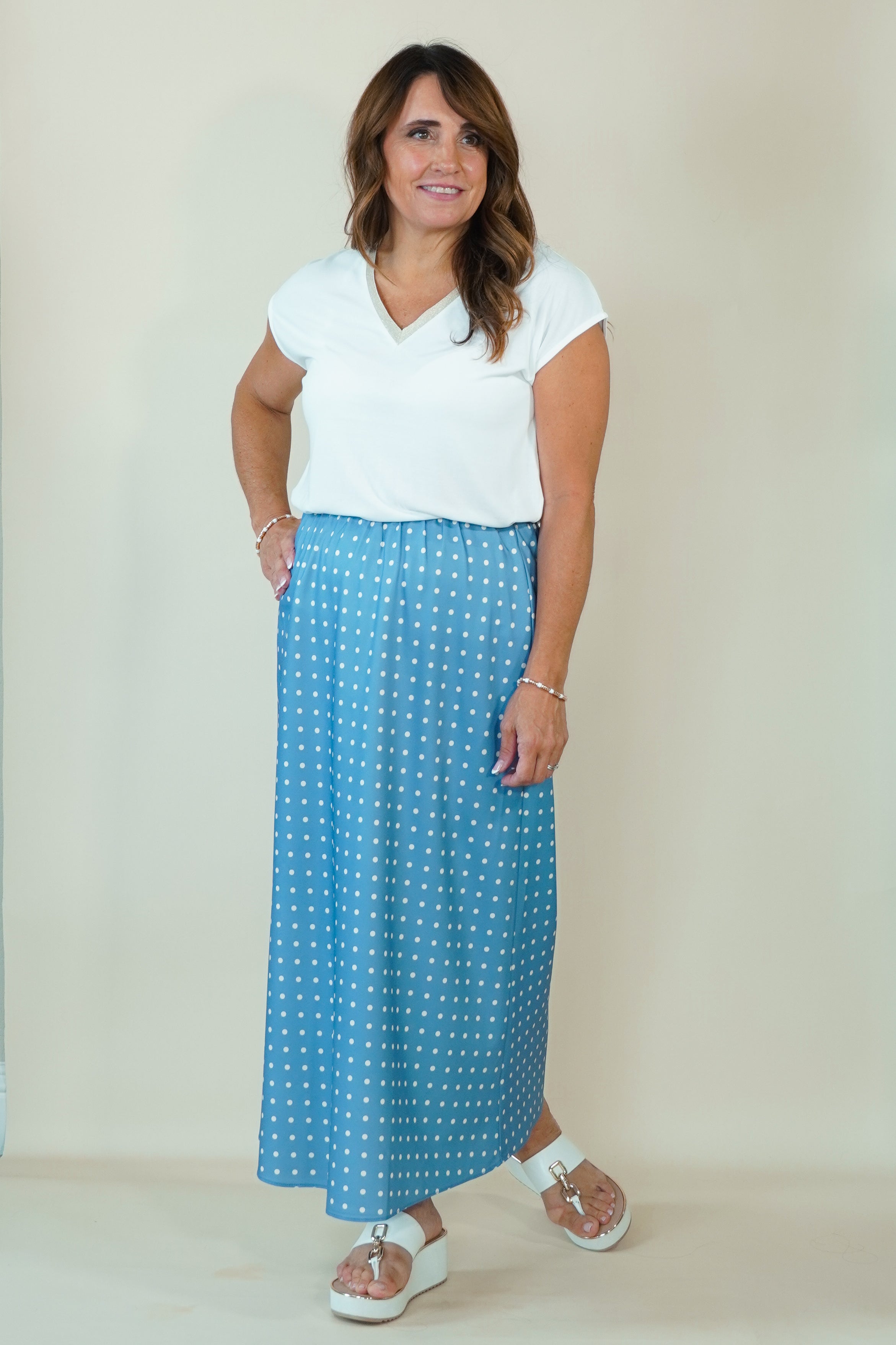 A woman modeling a white short-sleeve top and a blue maxi skirt with a white polka dot pattern, standing against a plain beige studio background.