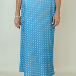 A woman modeling a white short-sleeve V-neck top and a light blue maxi skirt with a white polka dot pattern, standing against a plain beige studio background.