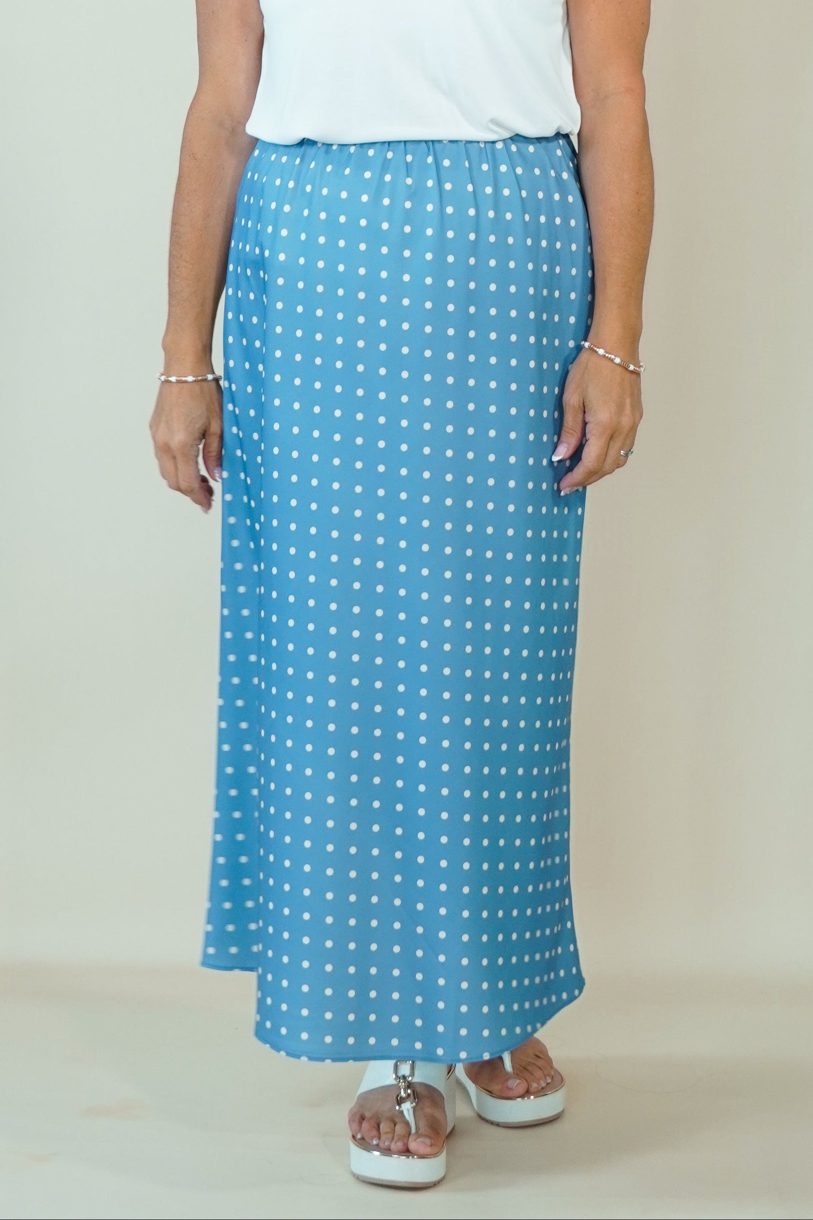 A woman modeling a white short-sleeve V-neck top and a light blue maxi skirt with a white polka dot pattern, standing against a plain beige studio background.