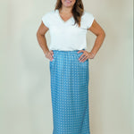 A woman modeling a white short-sleeve V-neck top and a blue maxi skirt with a white polka dot pattern, standing against a plain beige studio background.