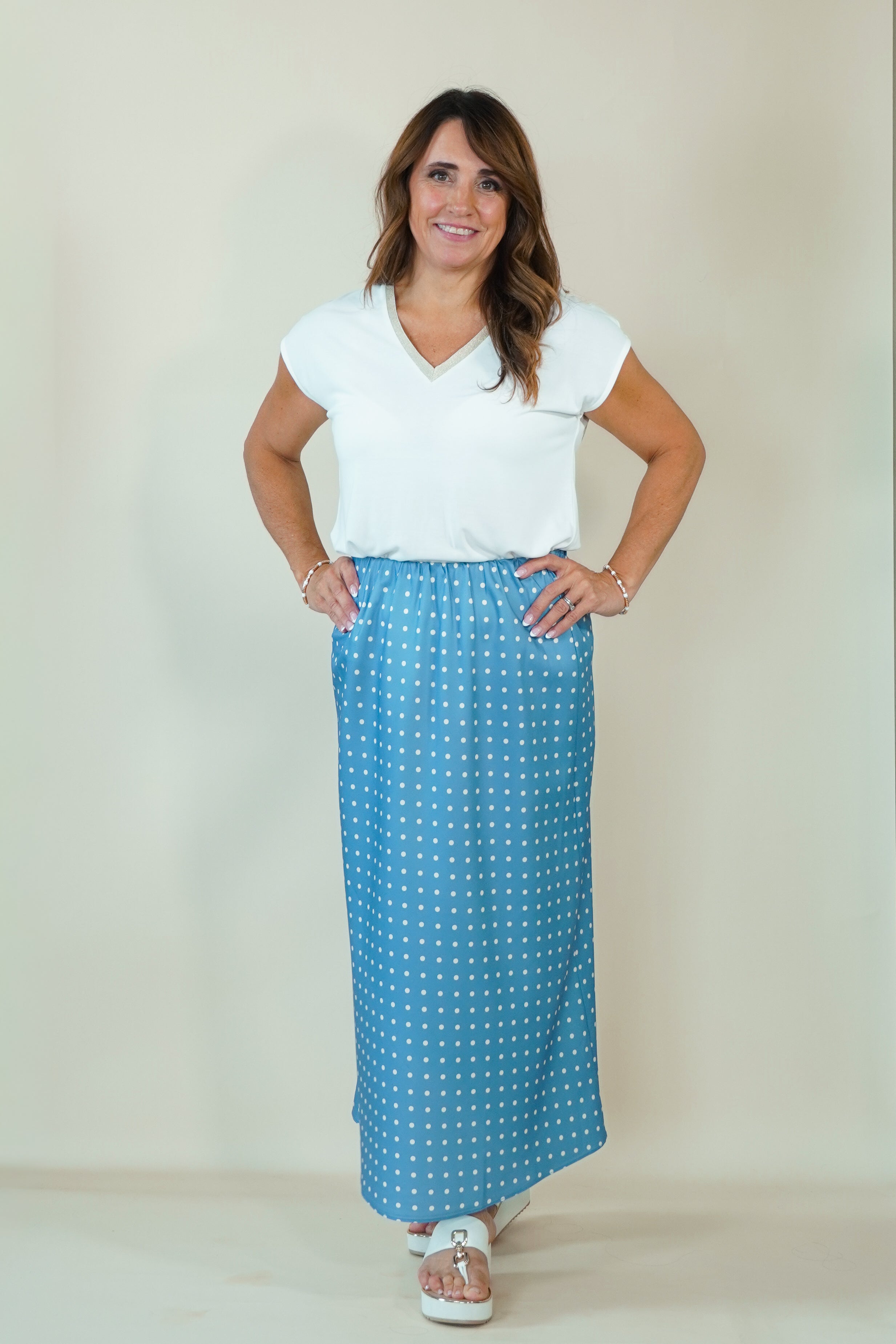 A woman modeling a white short-sleeve V-neck top and a blue maxi skirt with a white polka dot pattern, standing against a plain beige studio background.