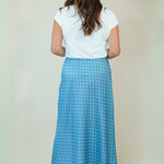 A woman viewed from behind wearing a white short-sleeve top and a blue midi skirt with a white polka dot pattern, standing against a plain beige studio background.