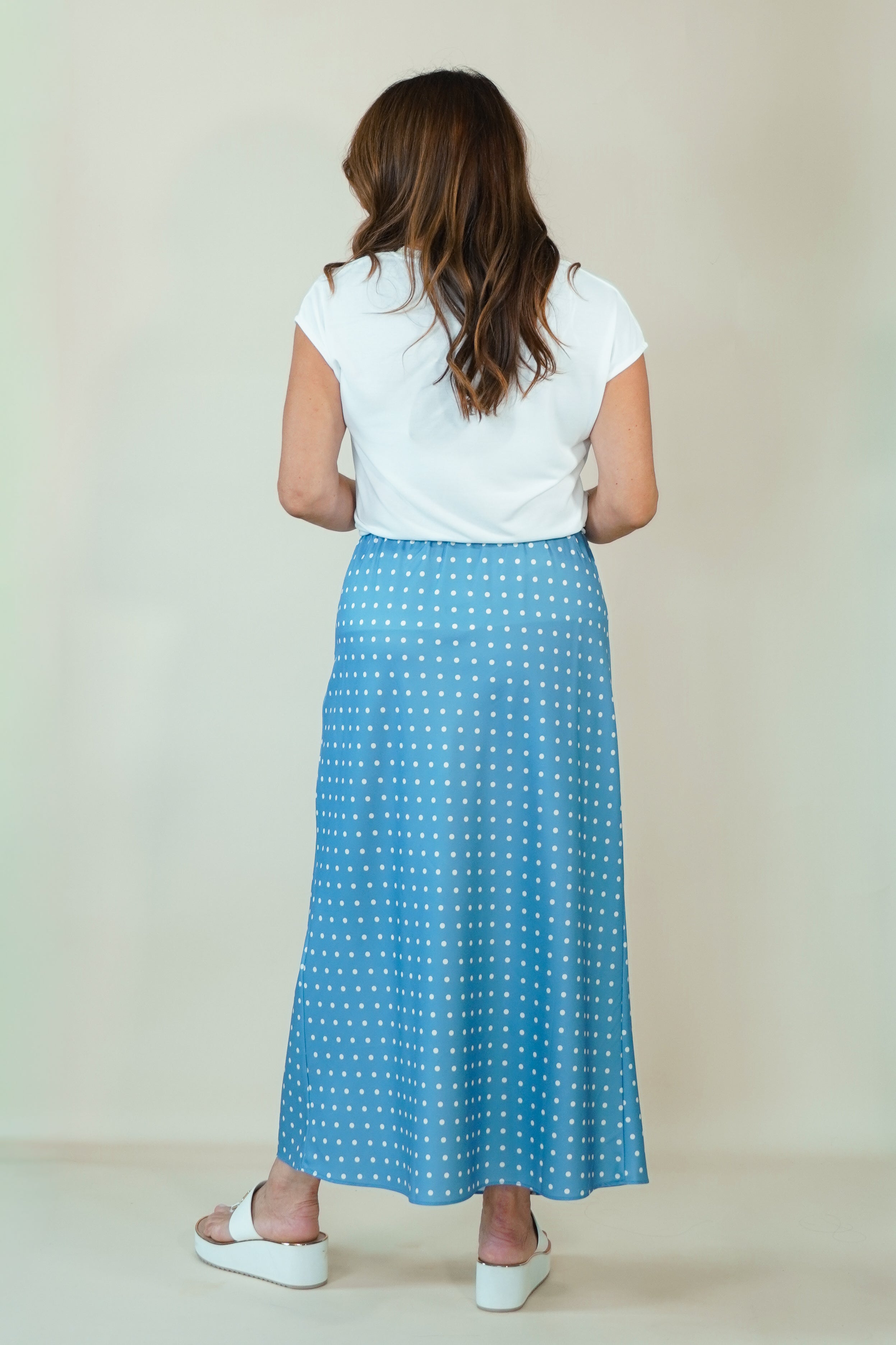 A woman viewed from behind wearing a white short-sleeve top and a blue midi skirt with a white polka dot pattern, standing against a plain beige studio background.