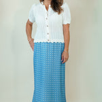 A woman modeling a two-piece outfit consisting of a white short-sleeve button-front knit top and a blue maxi skirt with a white polka-dot pattern, set against a plain beige studio background.