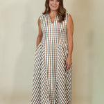 A woman modeling a sleeveless midi dress with a brown and beige gingham check pattern, standing against a plain beige studio backdrop.