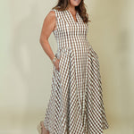 A woman modeling a sleeveless, V-neck maxi dress with a brown and cream gingham check pattern, standing against a solid light beige studio background.