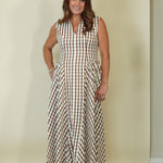 A woman modeling a sleeveless, V-neck maxi dress with a brown and beige gingham check pattern, standing against a plain beige studio backdrop.