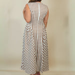 A woman viewed from behind wearing a sleeveless midi dress with a brown and white gingham check pattern, paired with beige wedge sandals, set against a plain beige studio background.