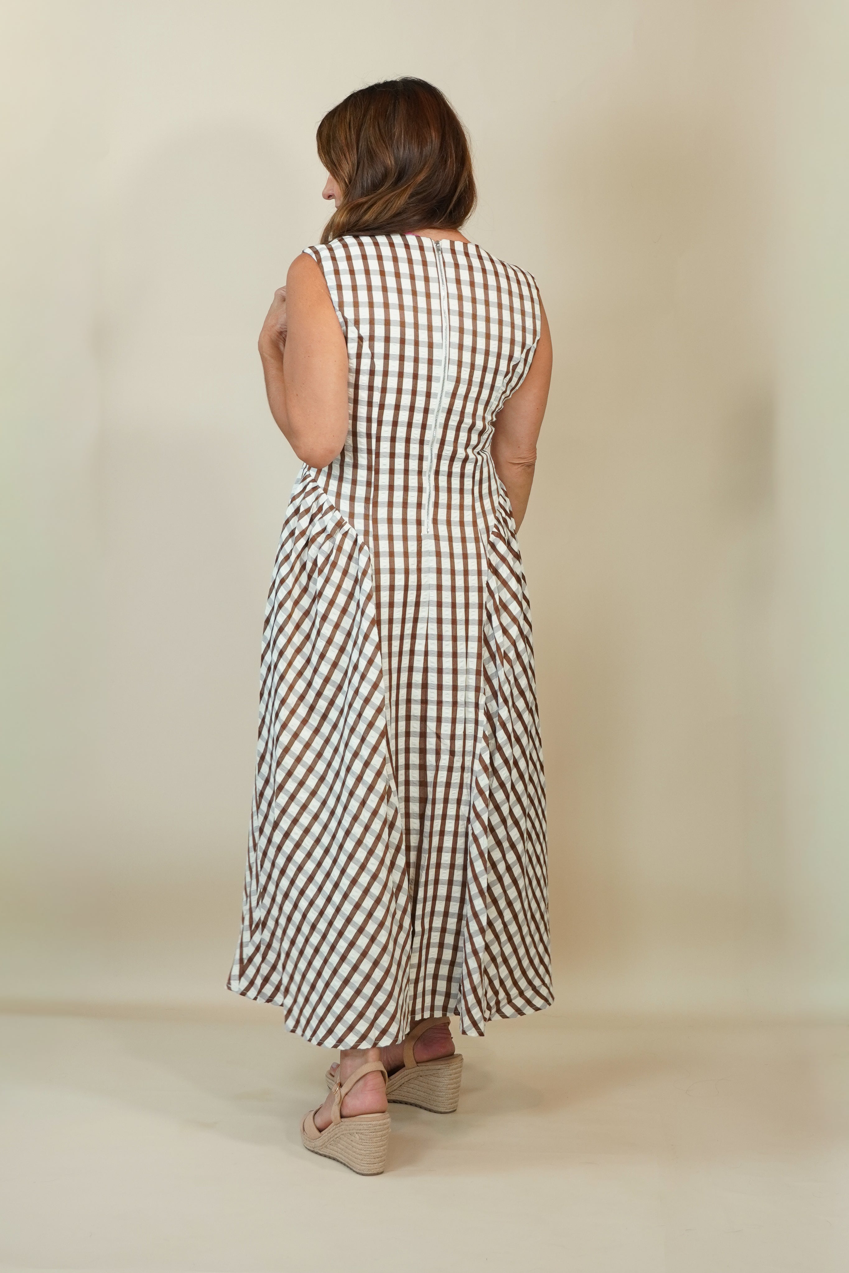 A woman viewed from behind wearing a sleeveless midi dress with a brown and white gingham check pattern, paired with beige wedge sandals, set against a plain beige studio background.