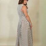 A woman modeling a sleeveless, knee-length gingham dress with a smocked bodice, shown from a side-back angle against a plain beige studio background.