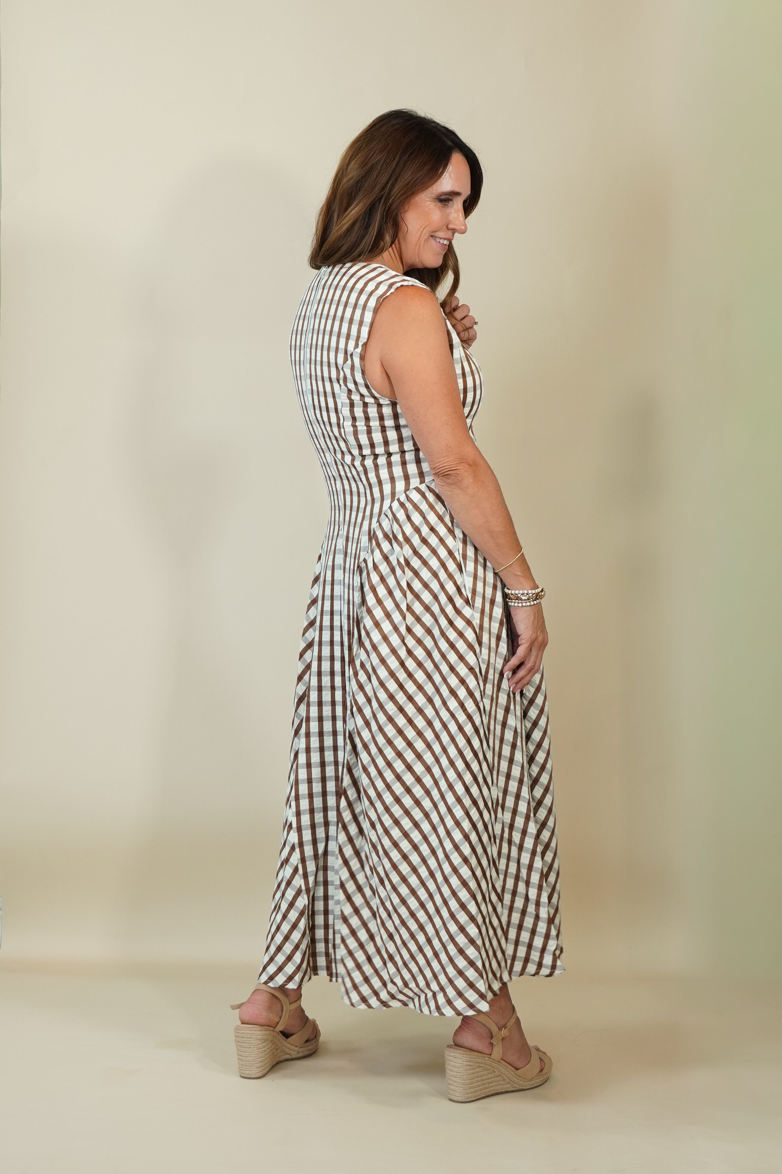 A woman modeling a sleeveless, knee-length gingham dress with a smocked bodice, shown from a side-back angle against a plain beige studio background.