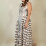 A woman modeling a sleeveless midi dress with a brown and white gingham check pattern, paired with beige wedge sandals, against a plain beige studio background.