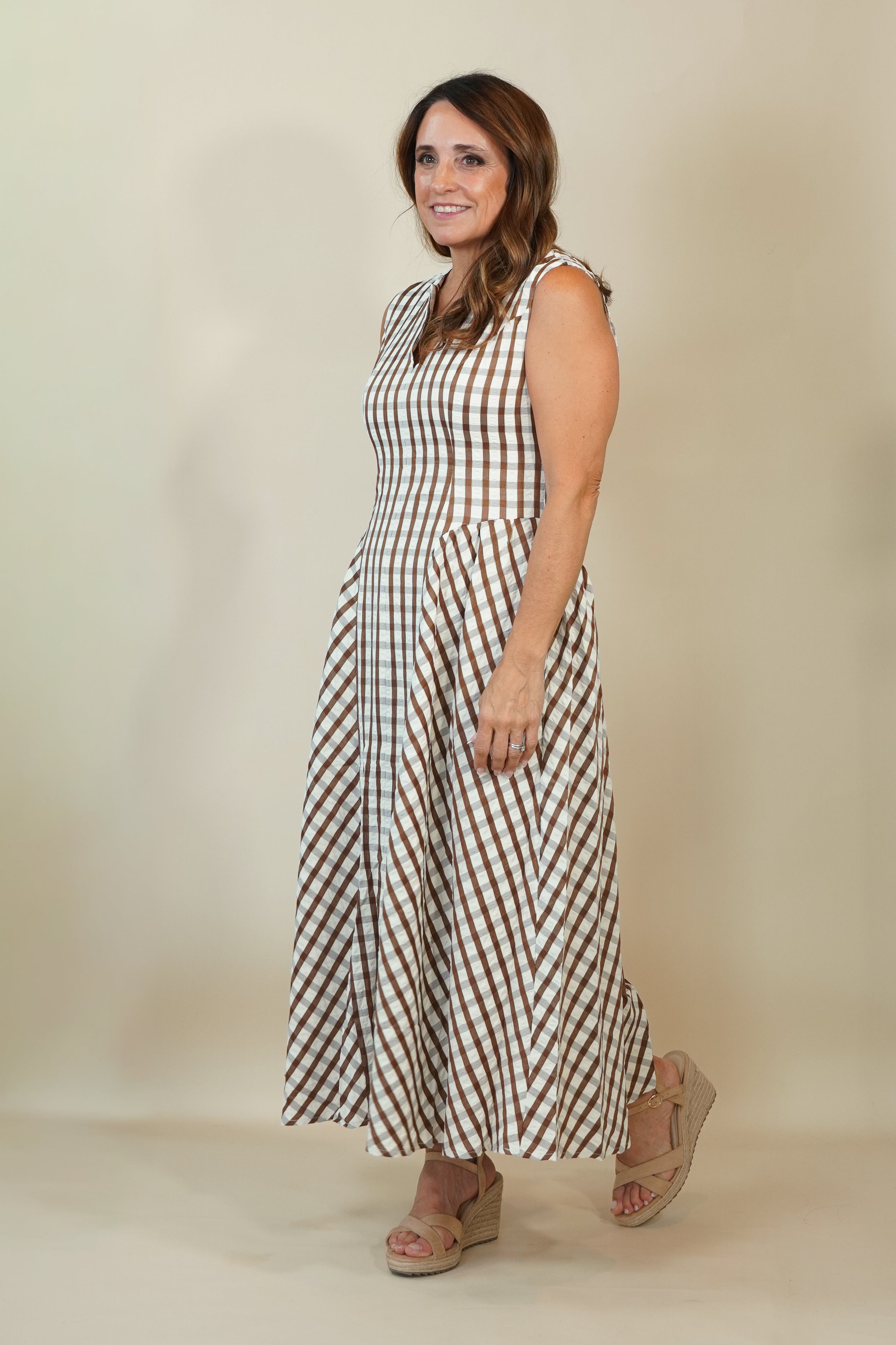 A woman modeling a sleeveless midi dress with a brown and white gingham check pattern, paired with beige wedge sandals, against a plain beige studio background.