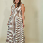 A woman modeling a sleeveless, V-neck maxi dress with a brown and cream gingham check pattern, standing against a plain beige studio background.