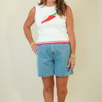 A woman modeling a sleeveless white knit top with a red and green chili pepper graphic on the chest and a red stripe at the hem, paired with light blue denim shorts, against a plain beige background.