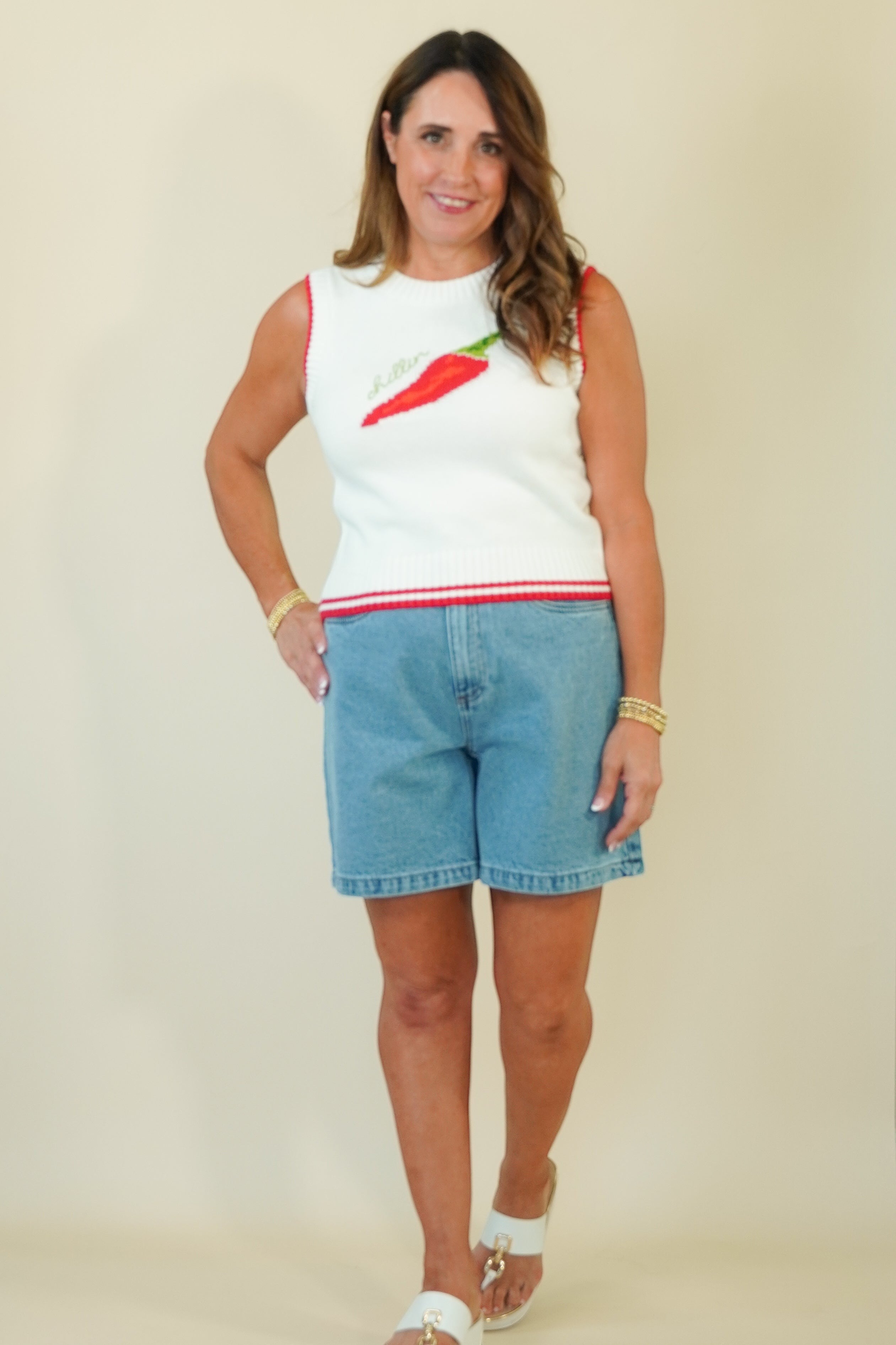 A woman modeling a sleeveless white knit top with a red and green chili pepper graphic on the chest and a red stripe at the hem, paired with light blue denim shorts, against a plain beige background.