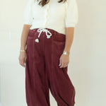 A woman modeling a pair of loose-fitting burgundy corduroy drawstring pants with a white short-sleeve top, standing against a plain white studio backdrop.
