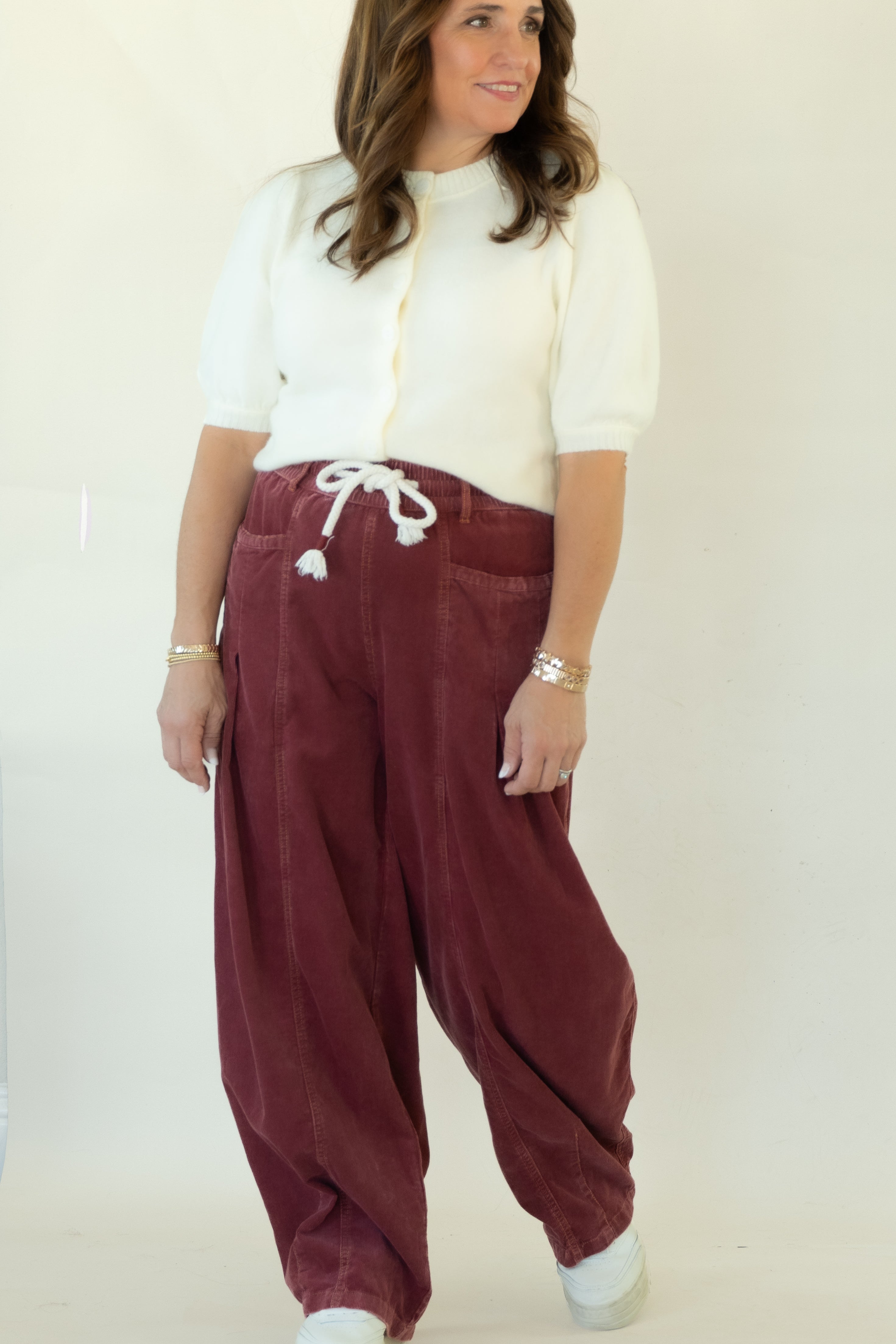 A woman modeling a pair of loose-fitting burgundy corduroy drawstring pants with a white short-sleeve top, standing against a plain white studio backdrop.