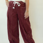 A woman modeling a pair of wide-leg burgundy corduroy pants with a white drawstring, paired with a white short-sleeve top and white sneakers, standing against a white studio backdrop.
