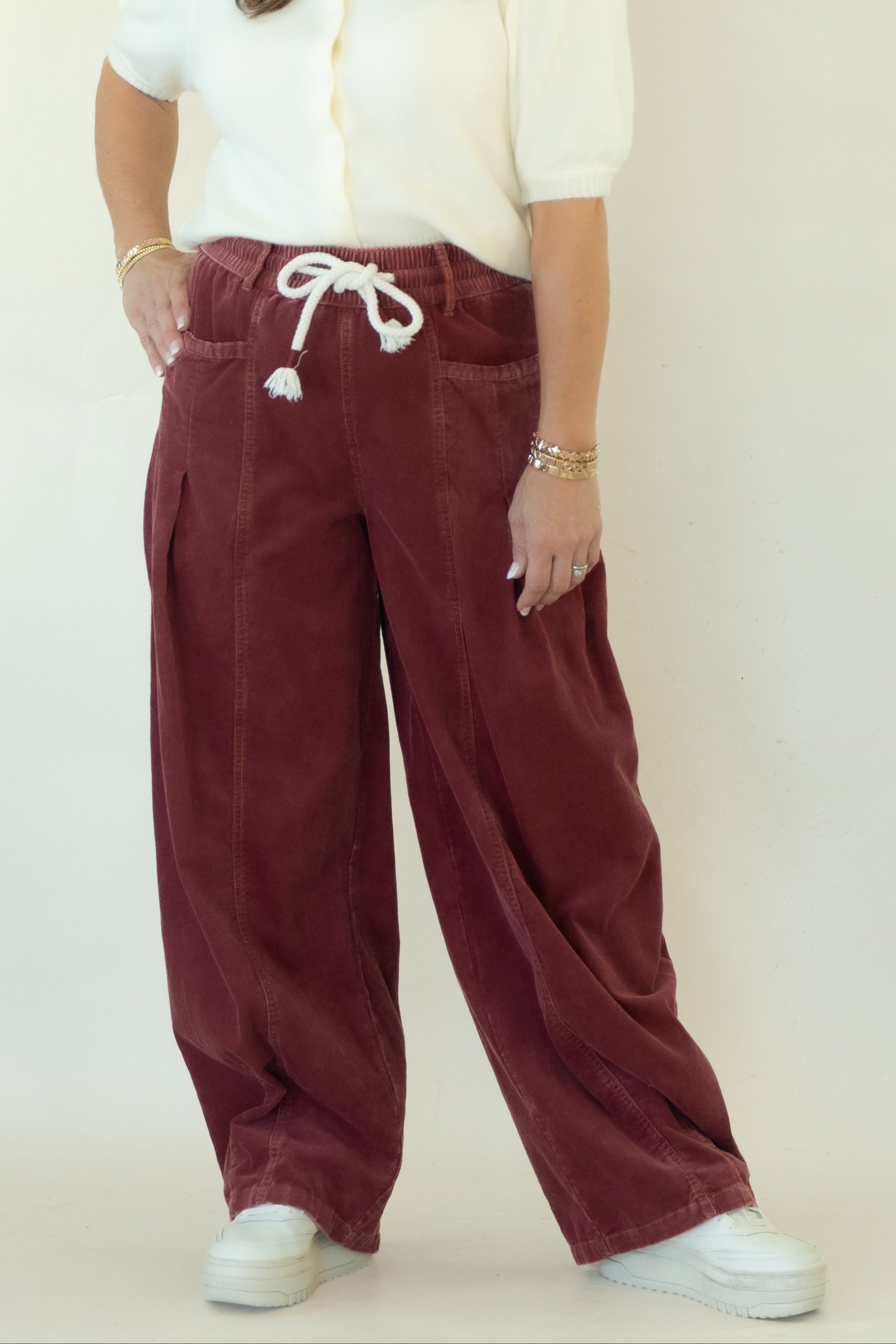 A woman modeling a pair of wide-leg burgundy corduroy pants with a white drawstring, paired with a white short-sleeve top and white sneakers, standing against a white studio backdrop.