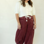 A woman modeling a pair of loose-fitting burgundy corduroy harem pants with a white drawstring, paired with a white short-sleeve top and white sneakers, standing against a white studio backdrop.