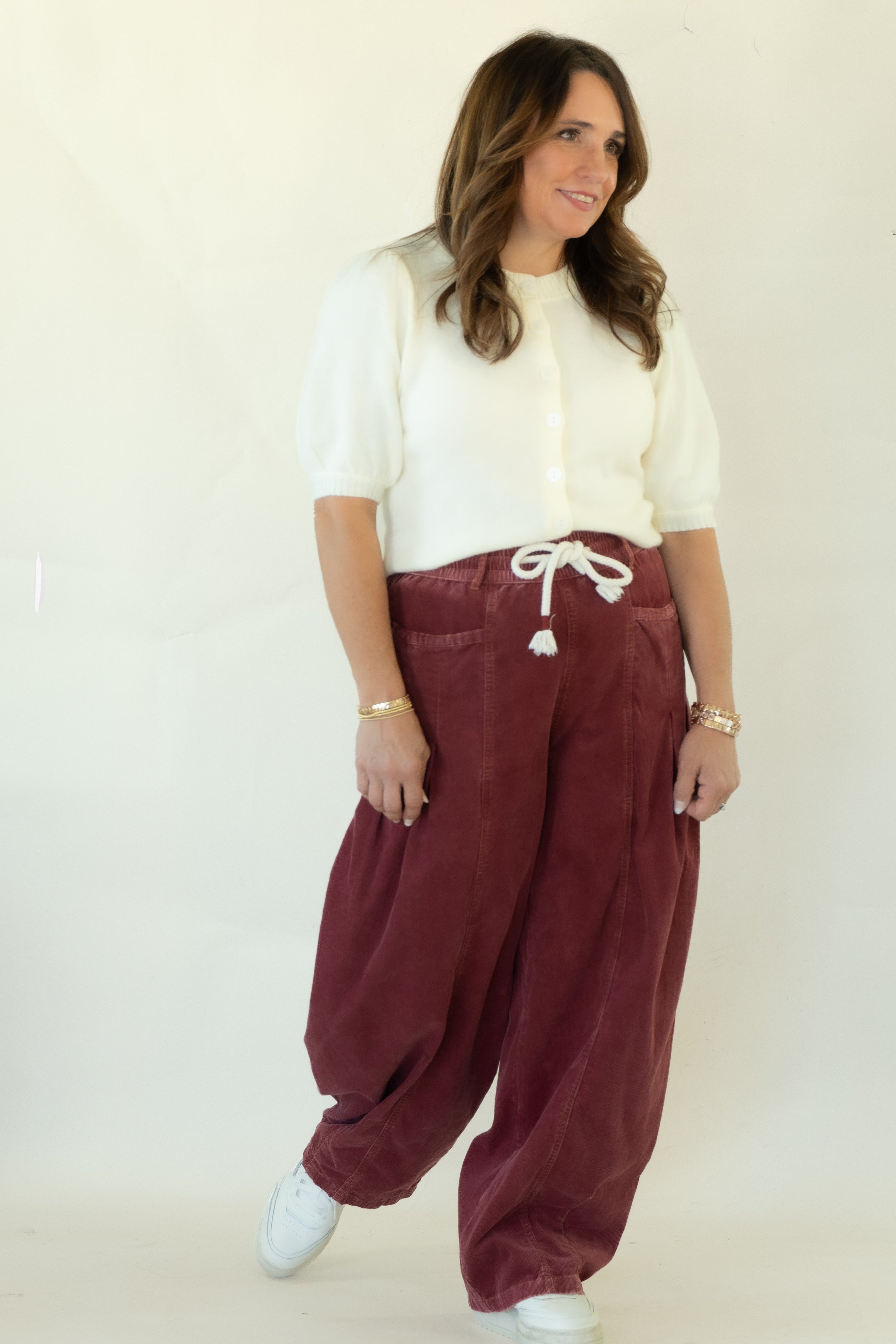 A woman modeling a pair of loose-fitting burgundy corduroy harem pants with a white drawstring, paired with a white short-sleeve top and white sneakers, standing against a white studio backdrop.