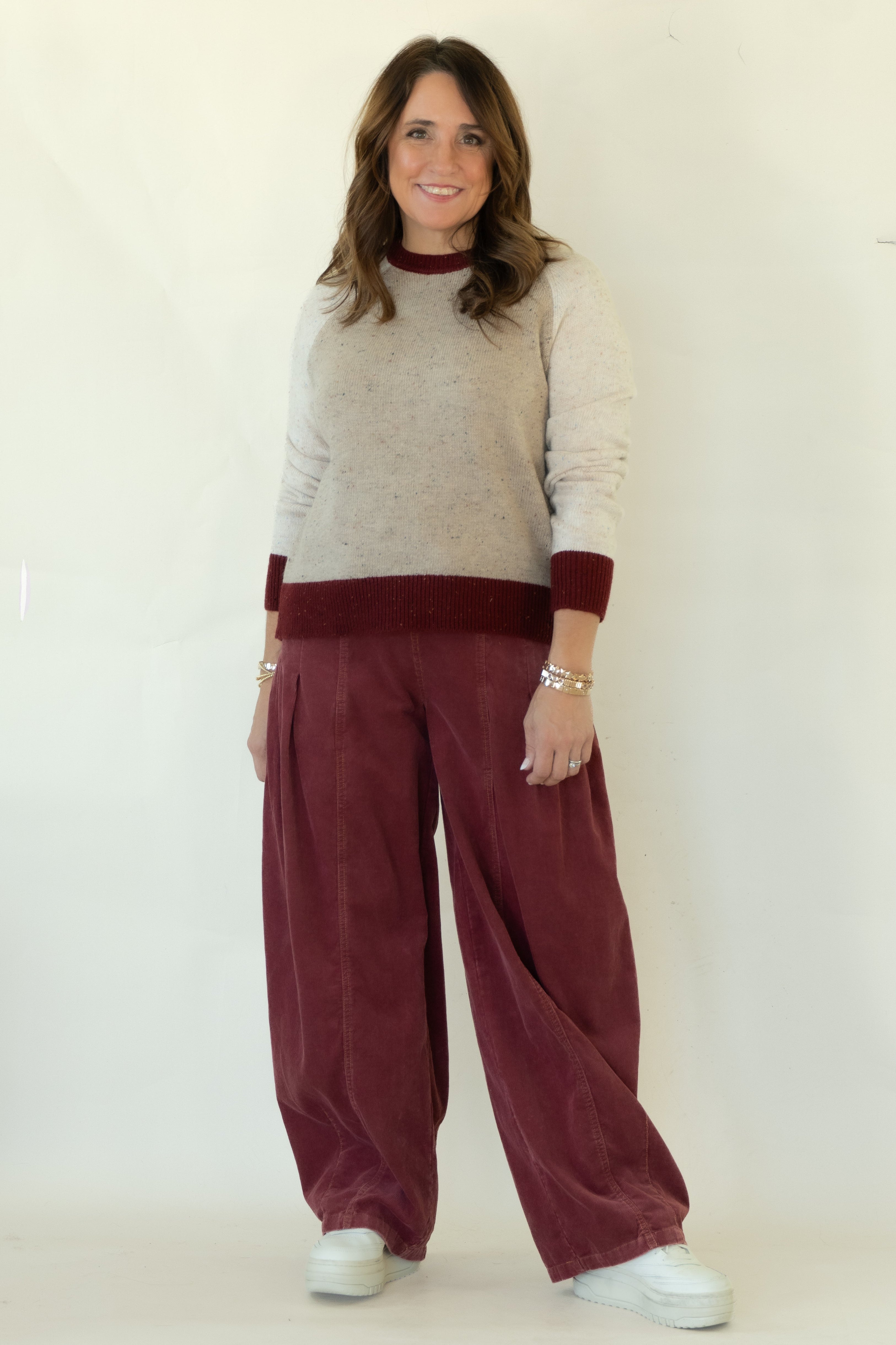 A woman modeling a knit crewneck sweater with a beige body and burgundy collar and cuffs, paired with wide-leg burgundy corduroy pants and white sneakers, standing against a white studio backdrop.