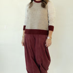 A woman modeling a knit sweater vest with cream and burgundy color blocking, paired with loose burgundy corduroy pants and white sneakers, set against a plain white studio backdrop.