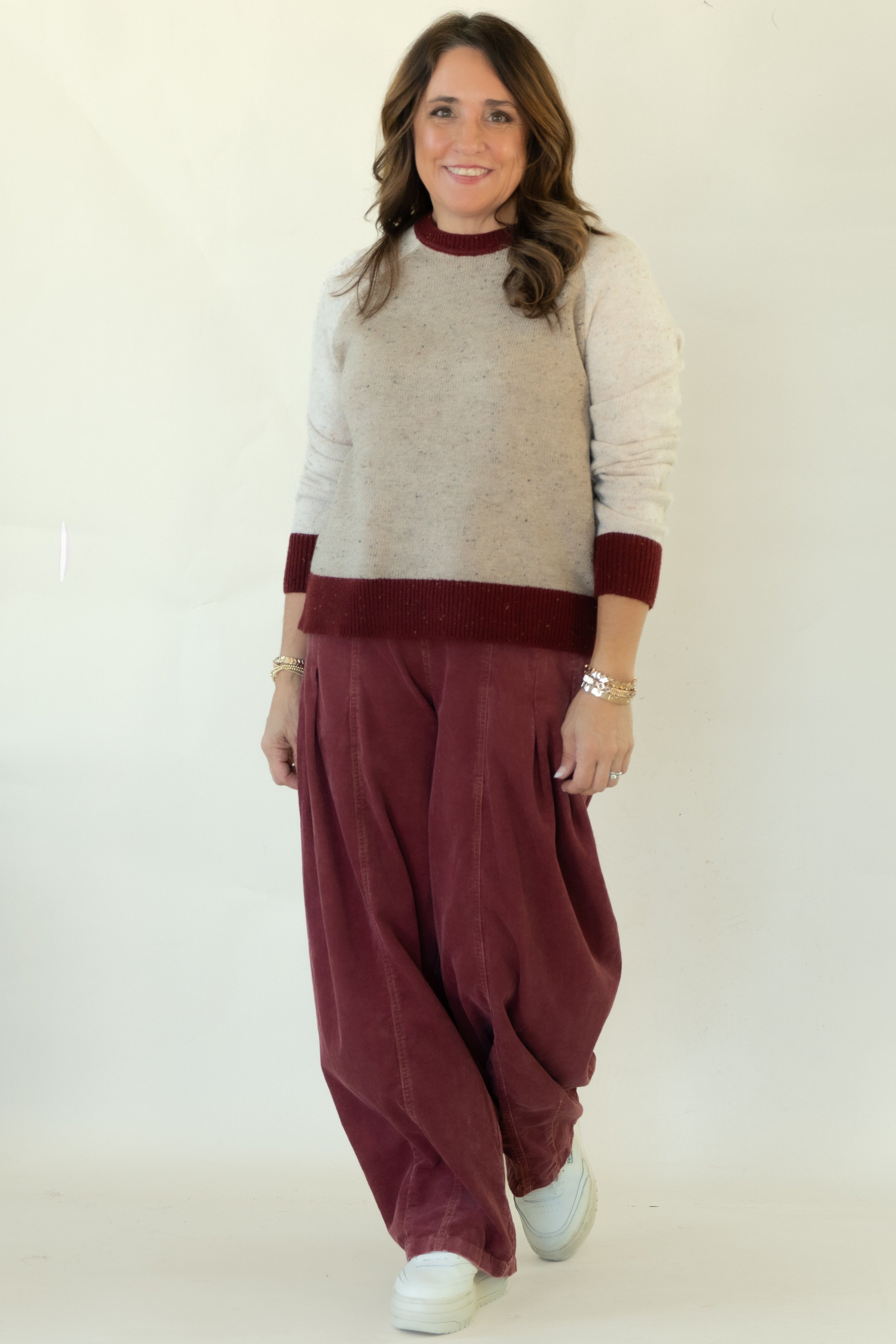 A woman modeling a knit sweater vest with cream and burgundy color blocking, paired with loose burgundy corduroy pants and white sneakers, set against a plain white studio backdrop.
