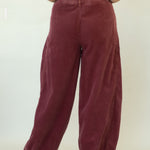 A woman viewed from behind wearing wide-leg burgundy corduroy trousers and a cream short-sleeve top, standing against a plain off-white studio backdrop.