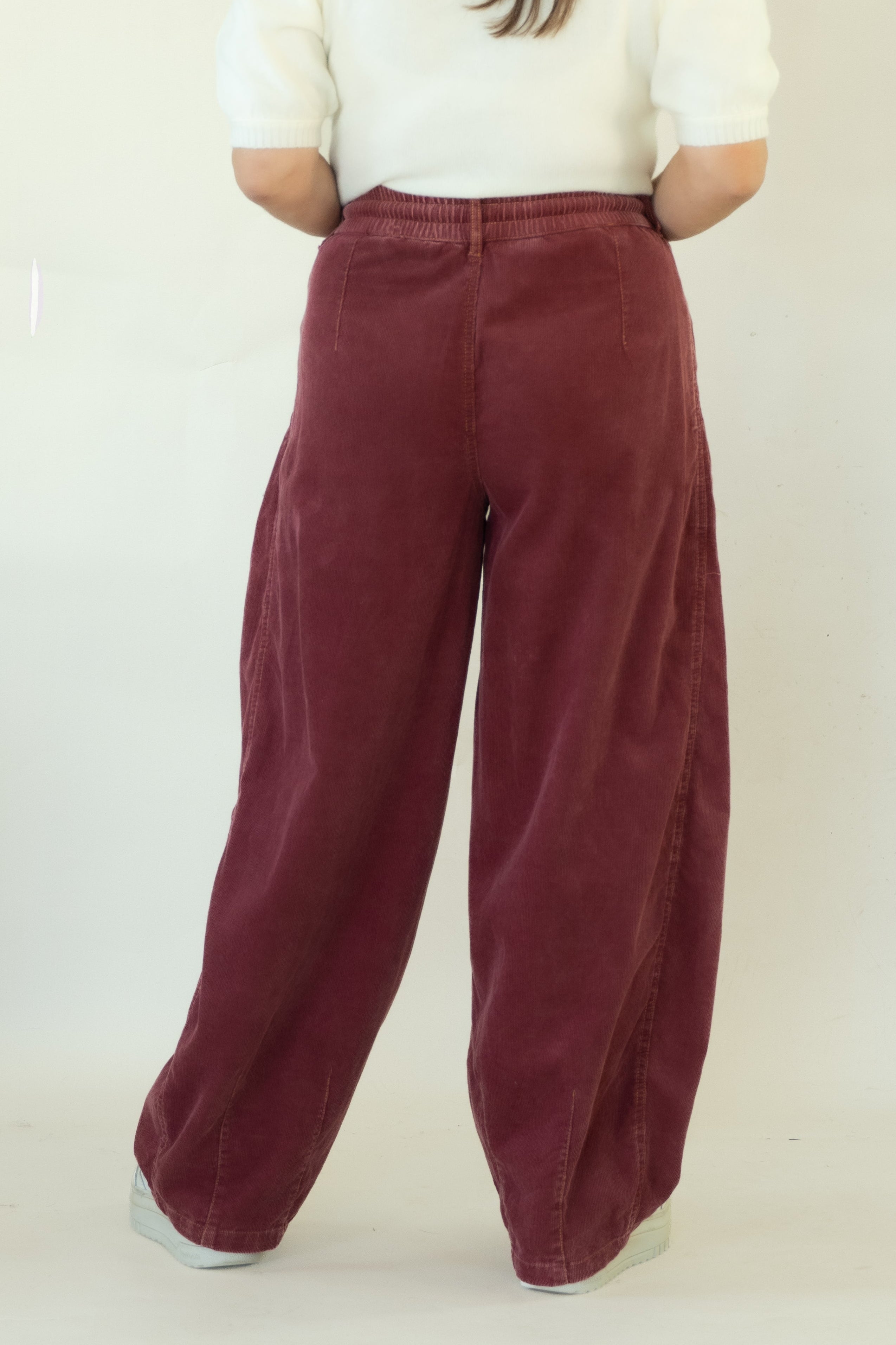 A woman viewed from behind wearing wide-leg burgundy corduroy trousers and a cream short-sleeve top, standing against a plain off-white studio backdrop.