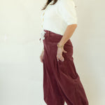 A woman modeling a pair of wide-leg burgundy corduroy pants with a white button-up shirt and white platform sneakers, standing against a plain white studio backdrop.