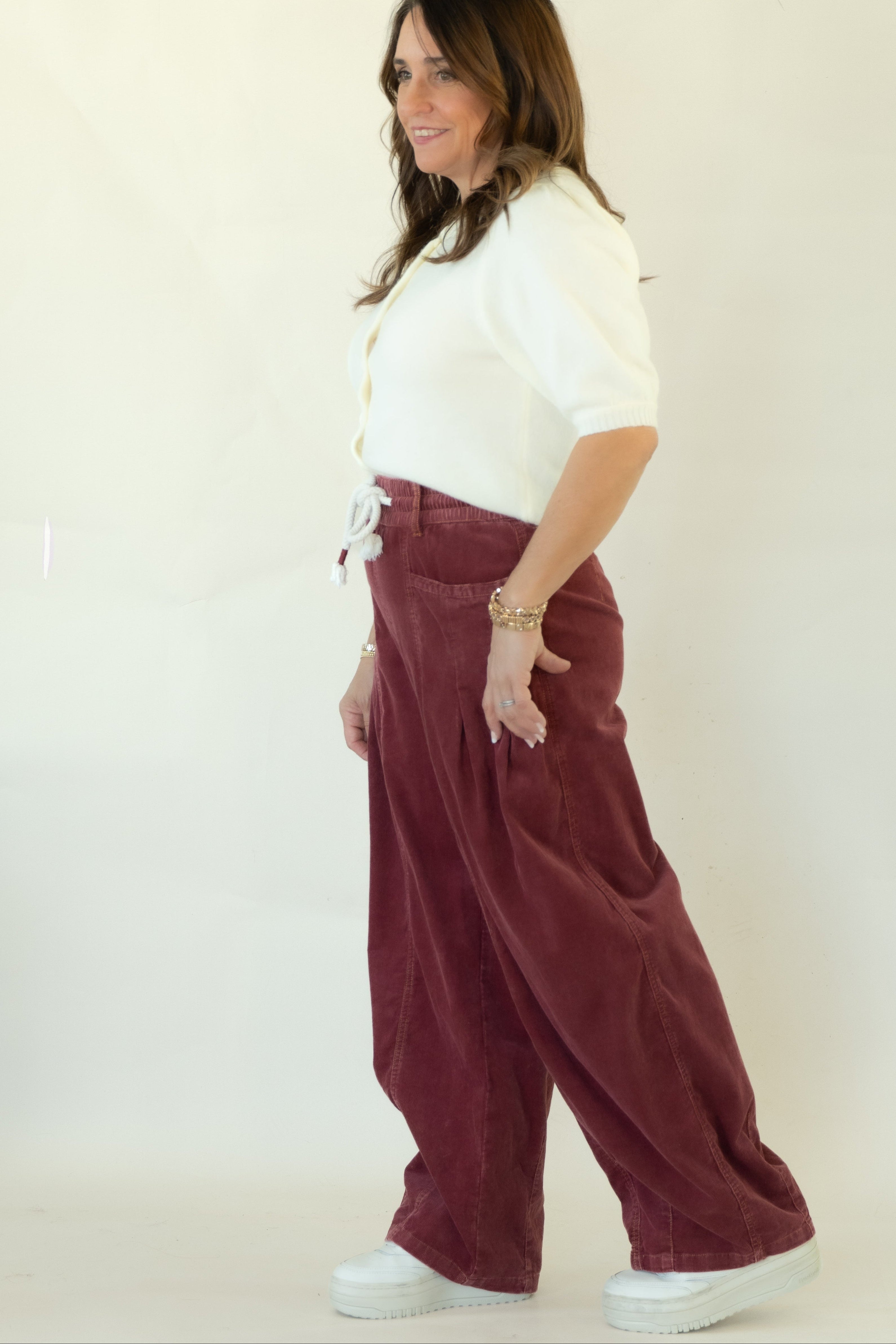 A woman modeling a pair of wide-leg burgundy corduroy pants with a white button-up shirt and white platform sneakers, standing against a plain white studio backdrop.