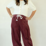 A woman modeling a pair of wide-leg burgundy corduroy pants with a white drawstring, paired with a white short-sleeve blouse and white sneakers, standing against a plain white studio backdrop.