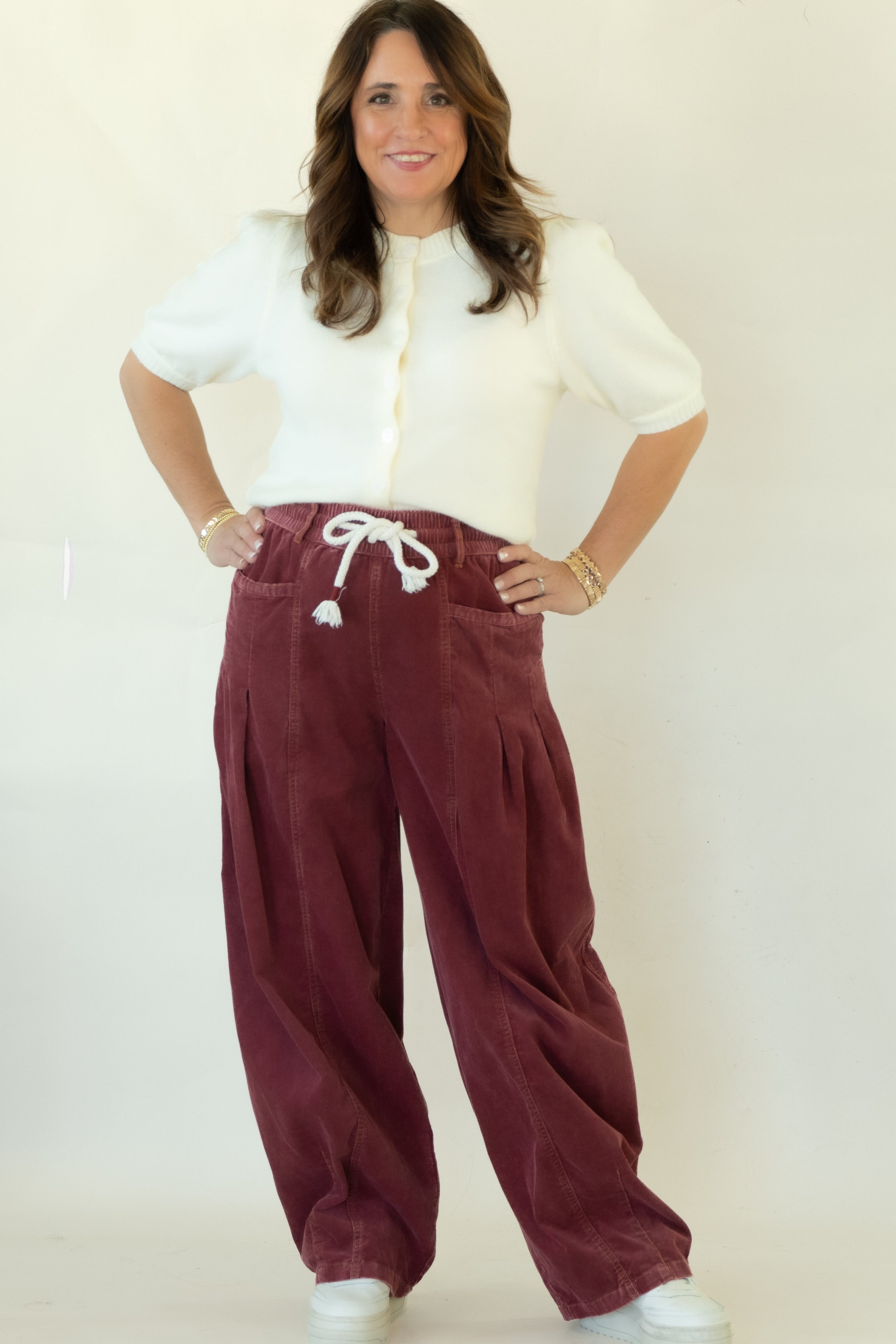 A woman modeling a pair of wide-leg burgundy corduroy pants with a white drawstring, paired with a white short-sleeve blouse and white sneakers, standing against a plain white studio backdrop.