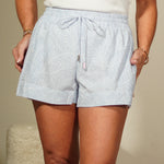 Woman in a white top and light blue shorts standing against a plain wall.