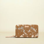 A zip-around wallet with a brown and white paint splatter pattern, featuring a small pink leather brand tab on the front and a matching pink zipper pull, set against a solid cream background.