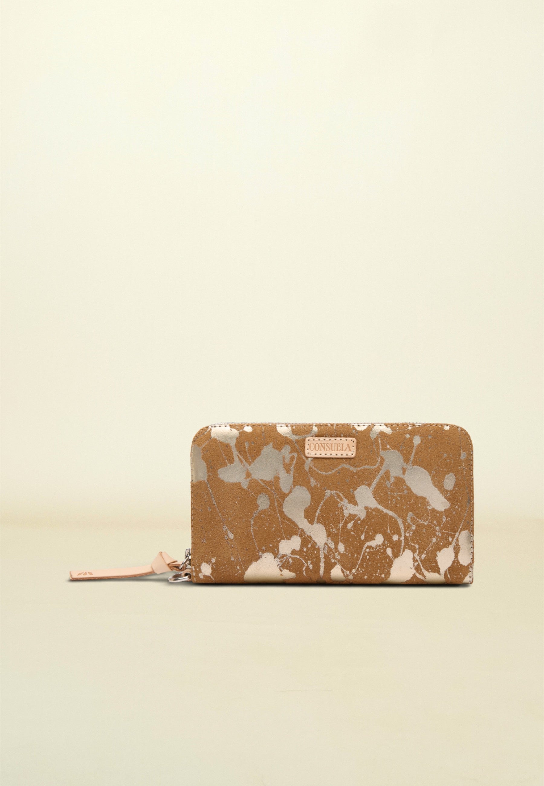 A zip-around wallet with a brown and white paint splatter pattern, featuring a small pink leather brand tab on the front and a matching pink zipper pull, set against a solid cream background.