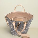 A structured bucket-style tote bag with a blue-gray and white abstract print exterior, light beige leather handles and shoulder strap, and a speckled beige interior, shown on a neutral light beige background.