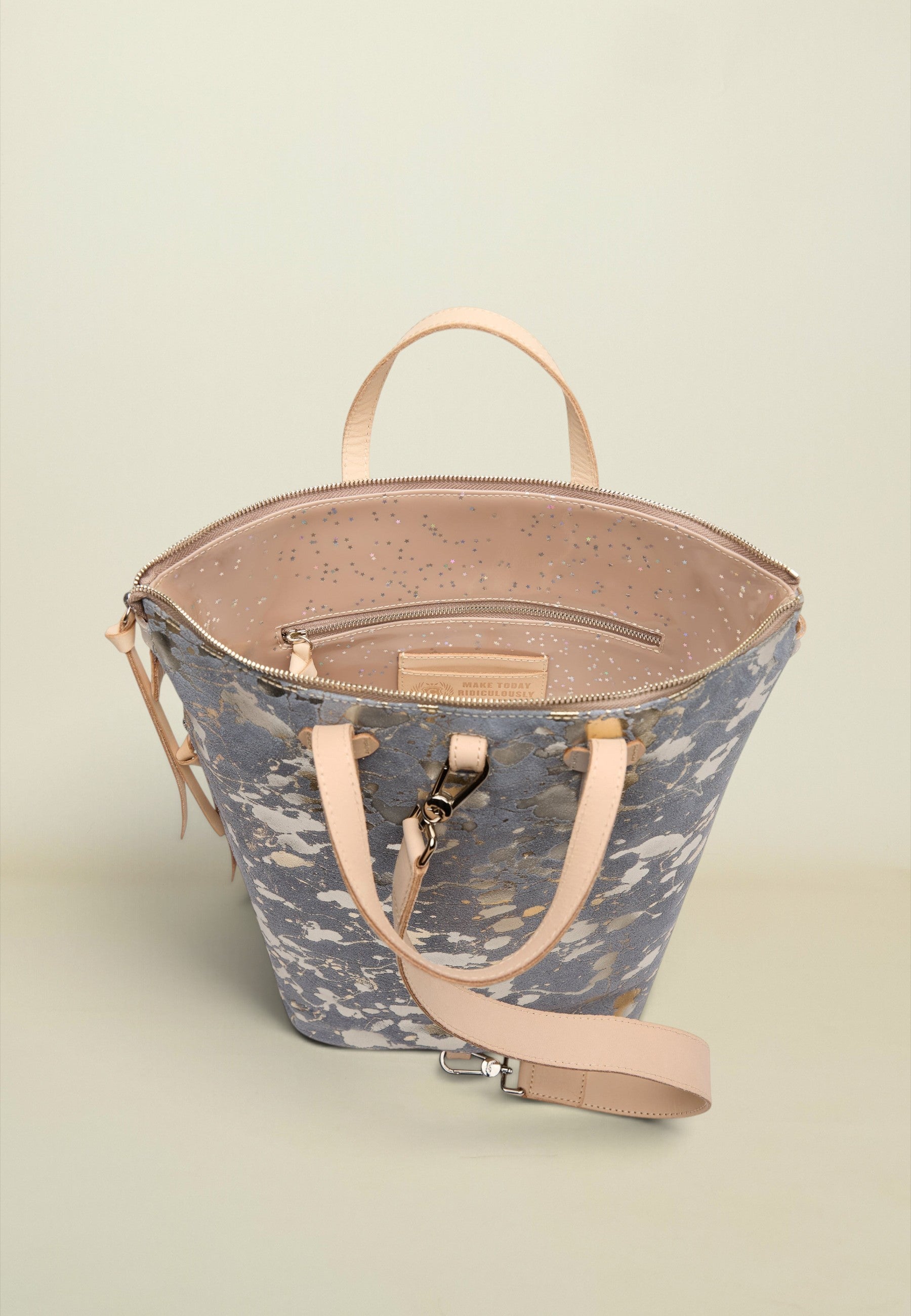 A structured bucket-style tote bag with a blue-gray and white abstract print exterior, light beige leather handles and shoulder strap, and a speckled beige interior, shown on a neutral light beige background.