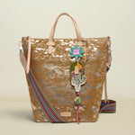 A gold-toned metallic tote bag with a distressed finish, featuring light beige leather handles, a colorful striped woven strap, and a decorative hanging charm with a flower and Hamsa hand.