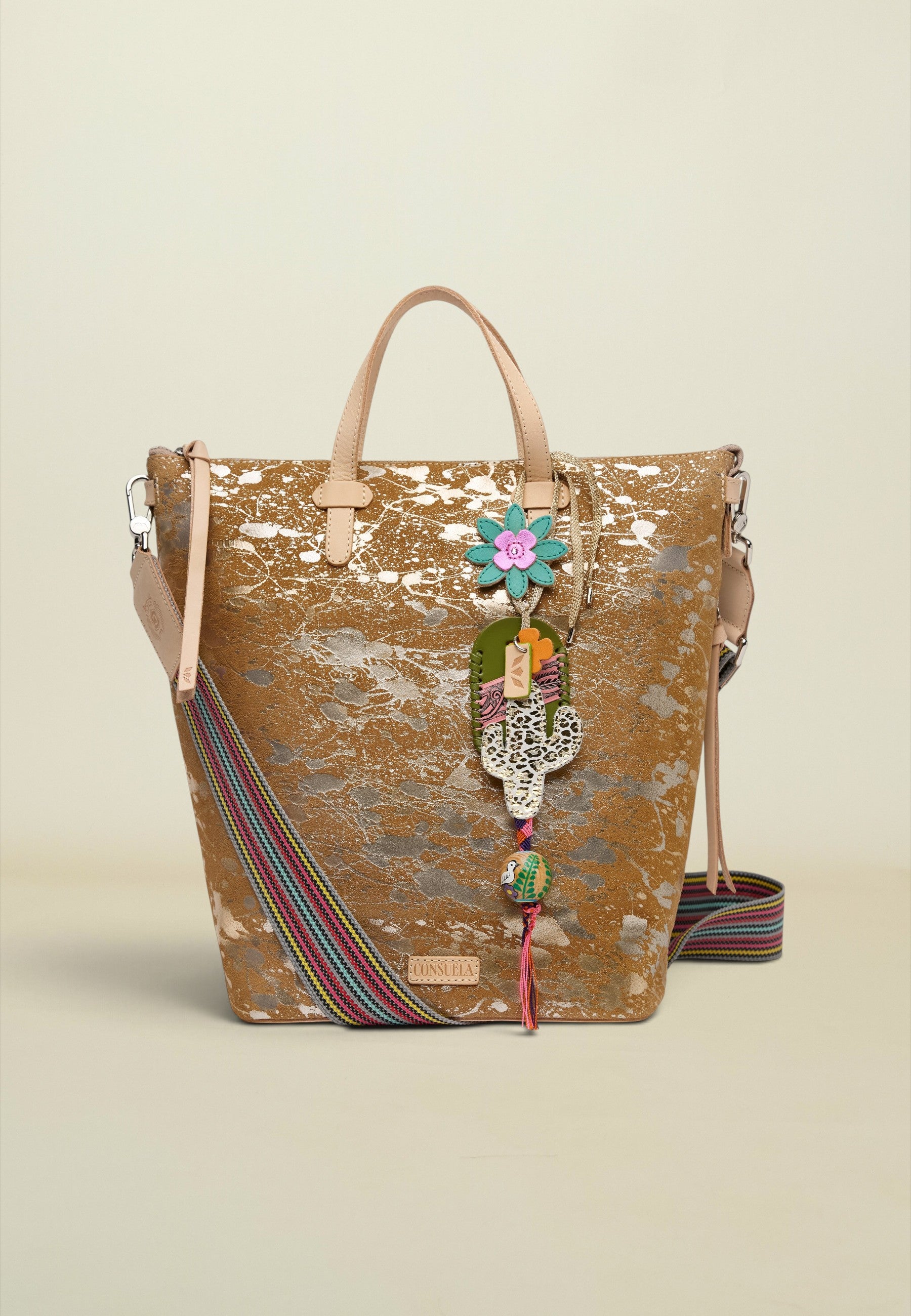 A gold-toned metallic tote bag with a distressed finish, featuring light beige leather handles, a colorful striped woven strap, and a decorative hanging charm with a flower and Hamsa hand.