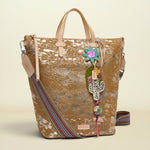 A bohemian-style tote bag with a distressed brown and beige pattern, featuring a colorful striped woven shoulder strap, a hanging Hamsa charm, and a flower accent, set against a solid cream background.
