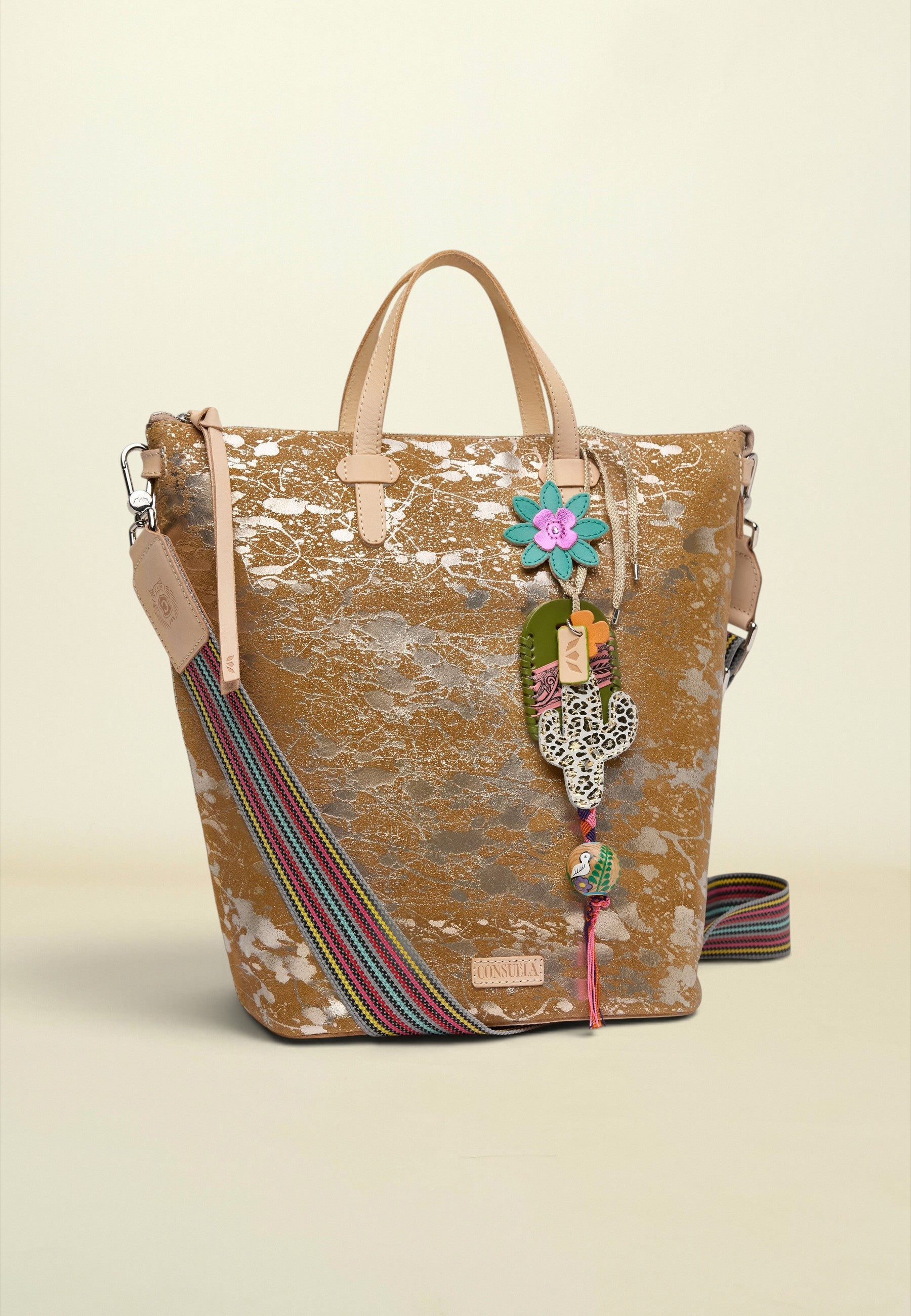 A bohemian-style tote bag with a distressed brown and beige pattern, featuring a colorful striped woven shoulder strap, a hanging Hamsa charm, and a flower accent, set against a solid cream background.