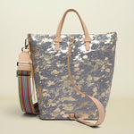 A metallic silver and gold marbled tote bag with light tan leather handles and a matching shoulder strap, shown next to a colorful striped detachable strap on a light beige background.