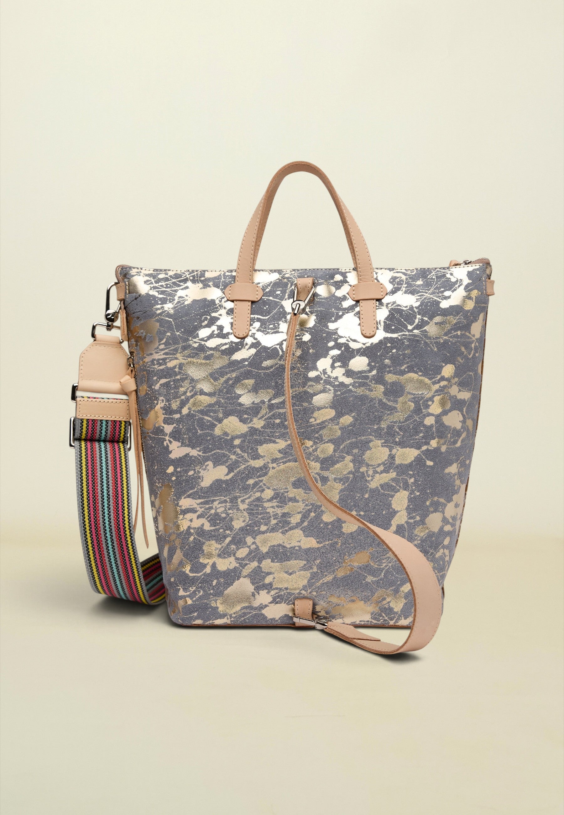 A metallic silver and gold marbled tote bag with light tan leather handles and a matching shoulder strap, shown next to a colorful striped detachable strap on a light beige background.