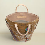 A Consuela handbag with a distressed brown and beige pattern, featuring light pink handles, a striped shoulder strap, and colorful embroidered details, set against a solid light beige background.